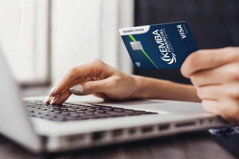 A woman holds a KEMBA Signature credit card while using a laptop, exemplifying the convenience of digital banking services.
