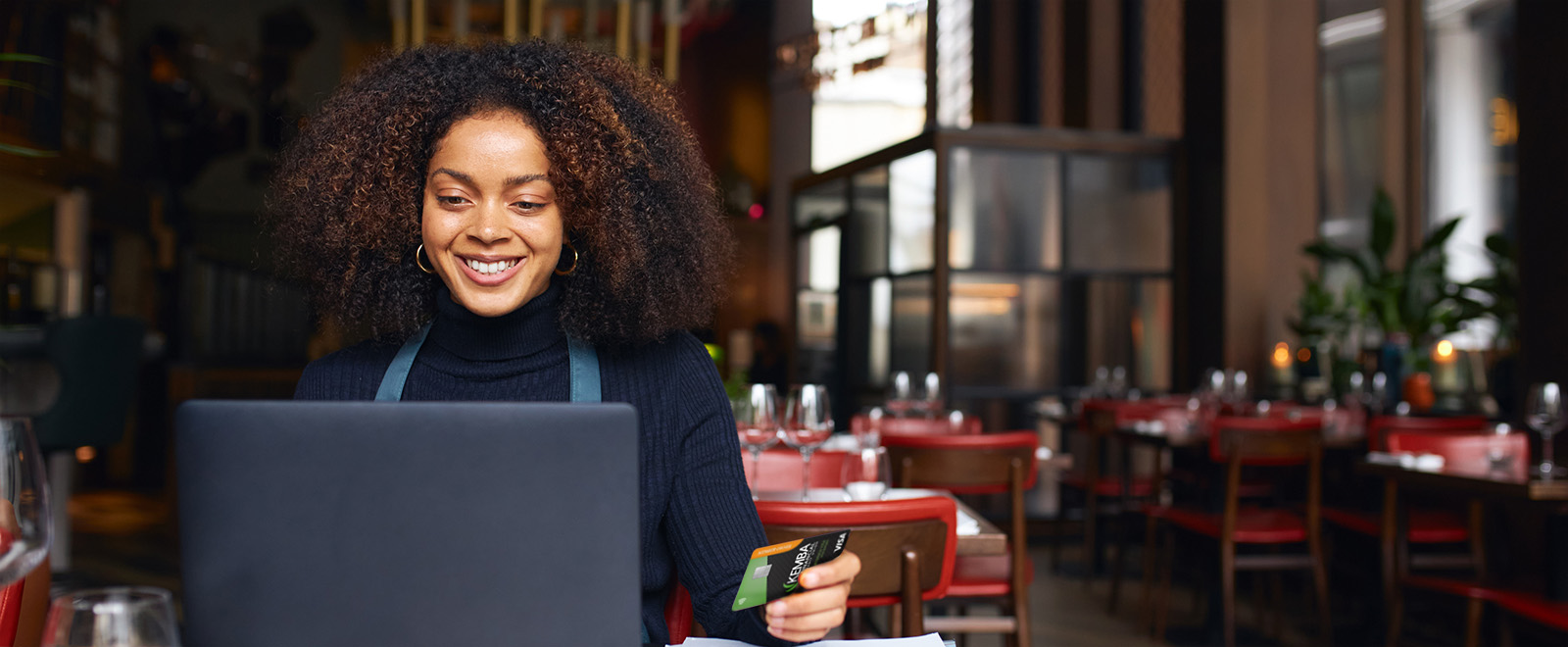 A woman sitting in a restaurant, smiling at her laptop while holding a credit card.