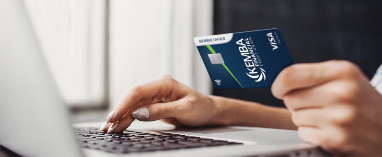 A woman holds a KEMBA Signature credit card while using a laptop, exemplifying the convenience of digital banking services.