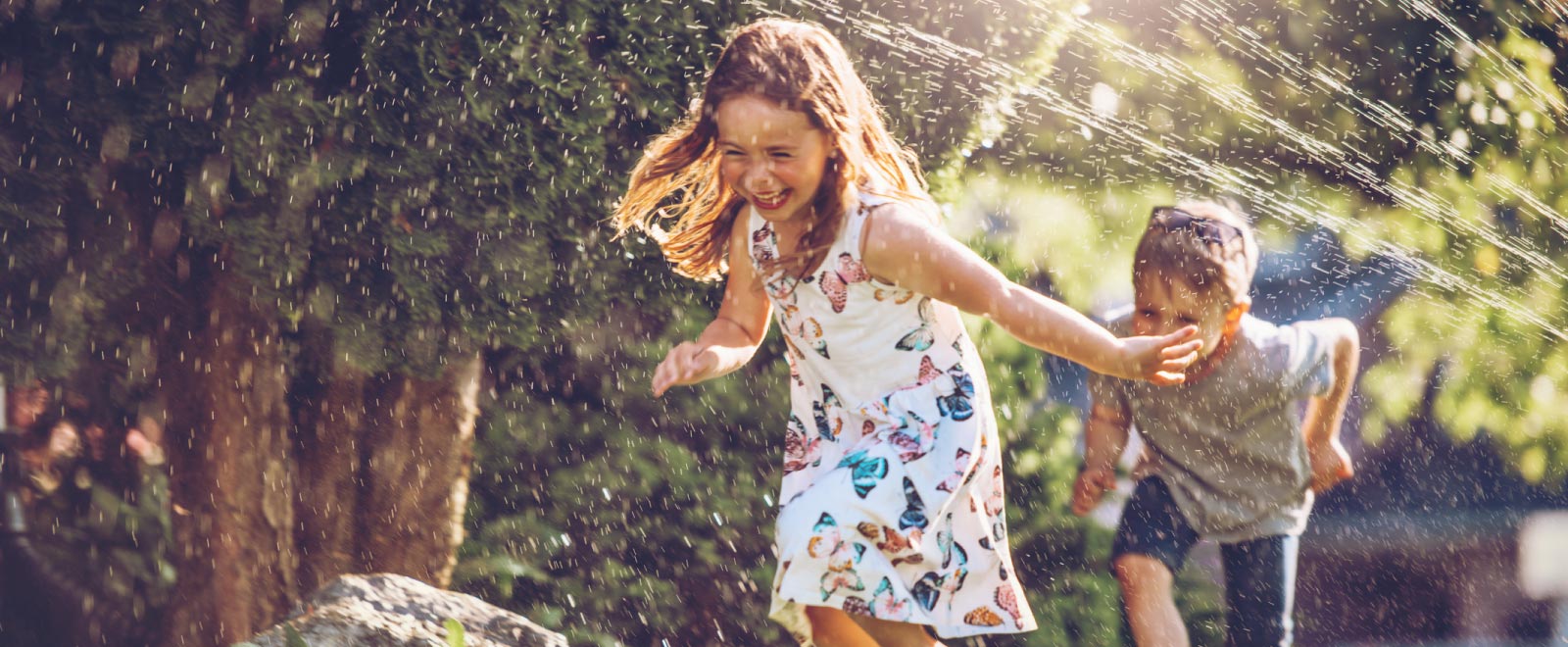 Two kids laughing and running under a sprinkler.